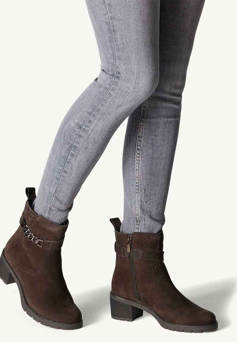 Bottines en suède marron avec un talon carré, fermeture éclair sur le côté et accent en chaîne ; associées à un jeans ajusté bleu clair. Texture lisse, design moderne.