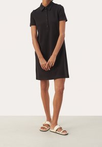 Persona che indossa un vestito nero a maniche corte con bottoni e sandali bianchi open-toe, in piedi con le mani leggermente incrociate davanti.