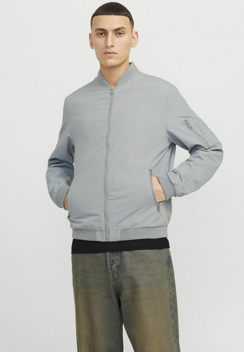 Veste bomber gris clair avec un devant zippé, des poignets et un ourlet côtelés, et des poches latérales. Texture lisse avec un design minimaliste.