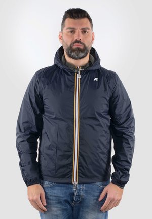 Giacca leggera blu navy con cappuccio, chiusura frontale a zip, polsini elastici e dettagli gialli a contrasto sulla zip. Materiale impermeabile con una consistenza liscia.