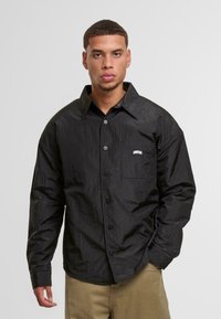 Fekete gombos ing, texturált anyagból, túlságosan bő szabású, hegyes gallérral és egy mellzsebkendővel, amelyen egy fehér "UNFAIR" logó található.