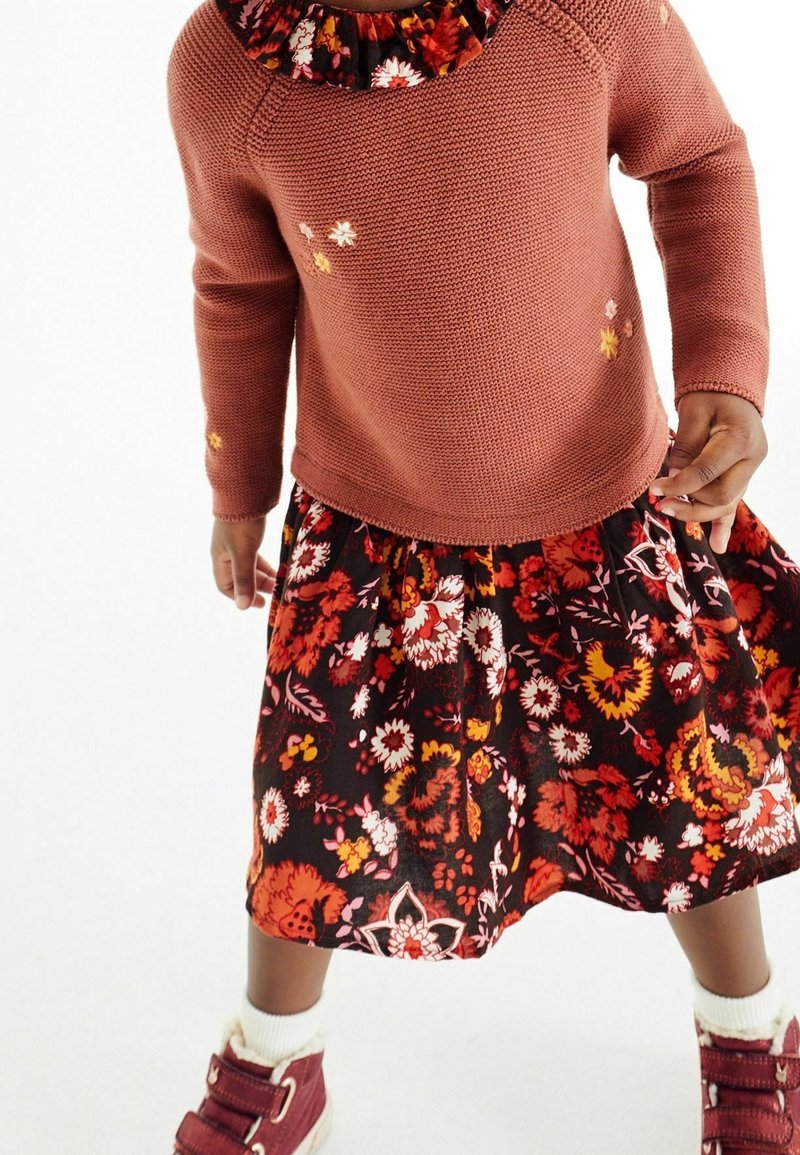 Enfant portant un pull en maille couleur rouille avec de petites fleurs brodées, une jupe à motif floral, des chaussettes blanches et des baskets montantes bordeaux.