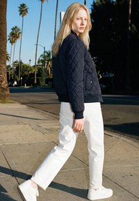 Donna bionda che cammina su un marciapiede assolato indossando una giacca trapuntata nera, pantaloni bianchi e sneakers bianche, con palme sullo sfondo.