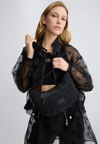 Zwarte stoffen tas met een textuurafwerking, ritscompartimenten en een verstelbare schouderband. Het model draagt een doorzichtige zwarte jas met een logopatroon.
