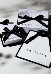 Trzy białe pudełka prezentowe z czarnymi wstążkami, z logo "LATELITA". Największe pudełko ma kształt kwadratu; wszystkie mają gładką, matową teksturę.