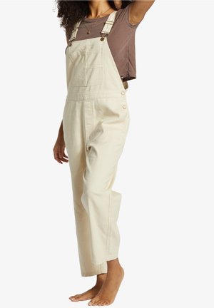 Kobieta ubrana w jasnobeżowe ogrodniczki i brązową koszulkę z krótkim rękawem, stojąca boso z jedną ręką uniesioną za głowę.