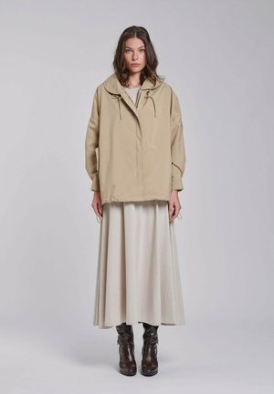 Donna con capelli castani fino alle spalle che indossa una giacca beige oversize, una lunga gonna crema e stivali marrone scuro al polpaccio, in piedi su uno sfondo chiaro.