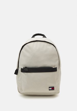 Beige backpack with black accents, a front zip pocket, and a top handle. Made from durable fabric, featuring a logo patch. Rounded shape.