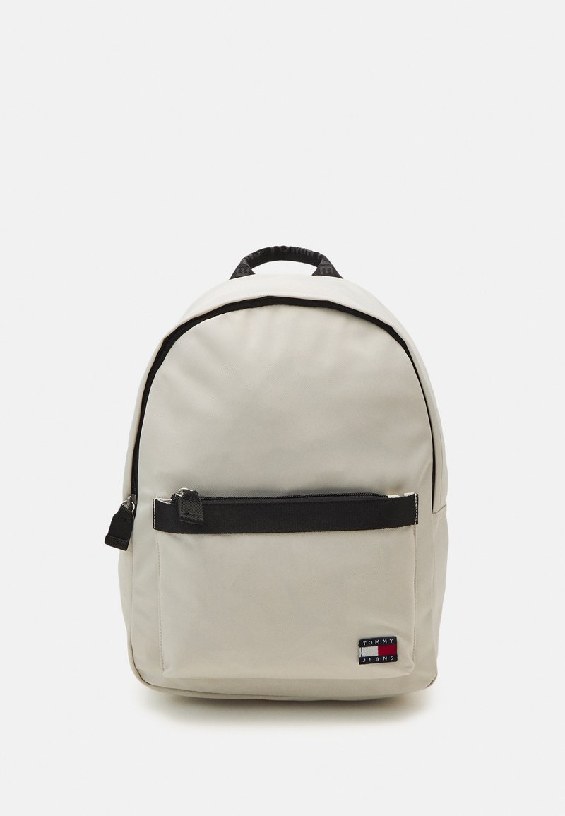 Sac à dos beige avec des accents noirs, une poche zippée à l'avant et une poignée en haut. Fait en tissu durable, avec un patch logo. Forme arrondie.