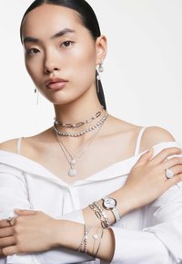 Srebrna biżuteria, w tym warstwowe naszyjniki, kolczyki, zegarek oraz pierścionki o okrągłych i owalnych kształtach, z gładkimi i teksturowanymi powierzchniami.