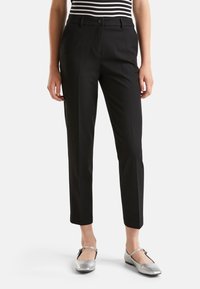 Vrouw in zwarte op maat gemaakte enkellange broek, gestreepte top en zilveren Mary Jane schoenen, staand tegen een witte achtergrond.