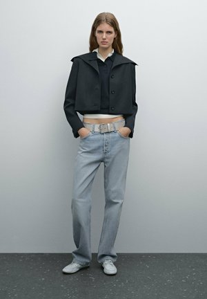 Veste noire raccourcie avec large col, associée à un polo sombre, un jean bleu clair et une ceinture grise. Le modèle se tient avec les mains dans les poches.