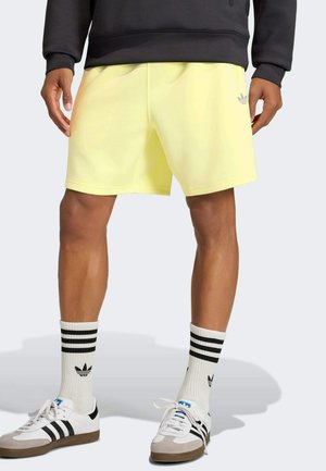 Person iført blege gule Adidas-shorts, hvide Adidas crew-sokker med sorte striber og hvide Adidas sneakers med sorte striber.