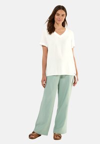 T-shirt bianco a V con maniche corte arrotolate, abbinato a pantaloni a gamba larga verde chiaro e sandali marroni con cinturini decorativi.