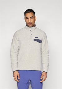 Pulover din fleece gri deschis, cu guler înalt, buzunar pe piept bleumarin și patru nasturi negri, având o textură moale și detalii în culoare albastră contrastantă.