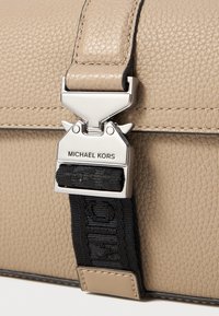 Kavos spalvos oda krepšys su tekstūruota paviršių, turintis sidabrinį metalinį užsegimą ir juodą diržą su "MICHAEL KORS" ženklu.