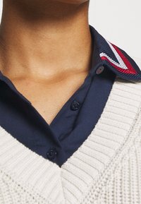 Krämfärgad stickad tröja med V-ringning över en mörkblå skjorta med krage. Kragen har röda, vita och blå randiga detaljer.