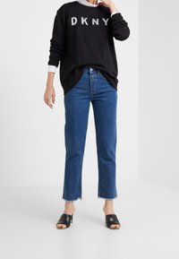 Person wearing black DKNY sweatshirt, blue cropped jeans with frayed hems, and black open-toe heeled mules standing in plain white space.