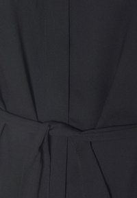 Gros plan d’un vêtement en tissu noir avec une ceinture nouée à la taille et des détails de couture visibles.