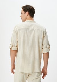 Camisa de color beige claro, de manga corta, con rayas verticales, puños abotonados y un corte relajado. La textura de la tela parece suave y transpirable.