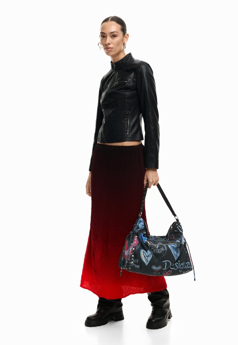 Žena v černé kožené bundě, červené sukní s ombre efektem, černých botách, držící černou kabelku s barevnými srdíčky a textovými motivy.