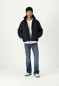 Μαύρο φουσκωτό μπουφάν με ψηλό γιακά, κλείσιμο με φερμουάρ και ριγωτούς μανσέτες, σε συνδυασμό με ανοιχτό μπλε τζιν ίσια γραμμή και λευκά αθλητικά παπούτσια.