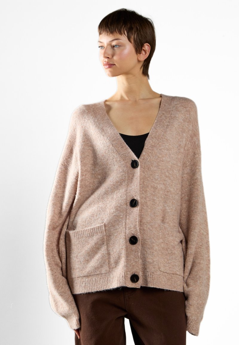 Cardigan oversize rosa chiaro in tessuto morbido, con scollo a V profondo, cinque bottoni neri e una tasca frontale.