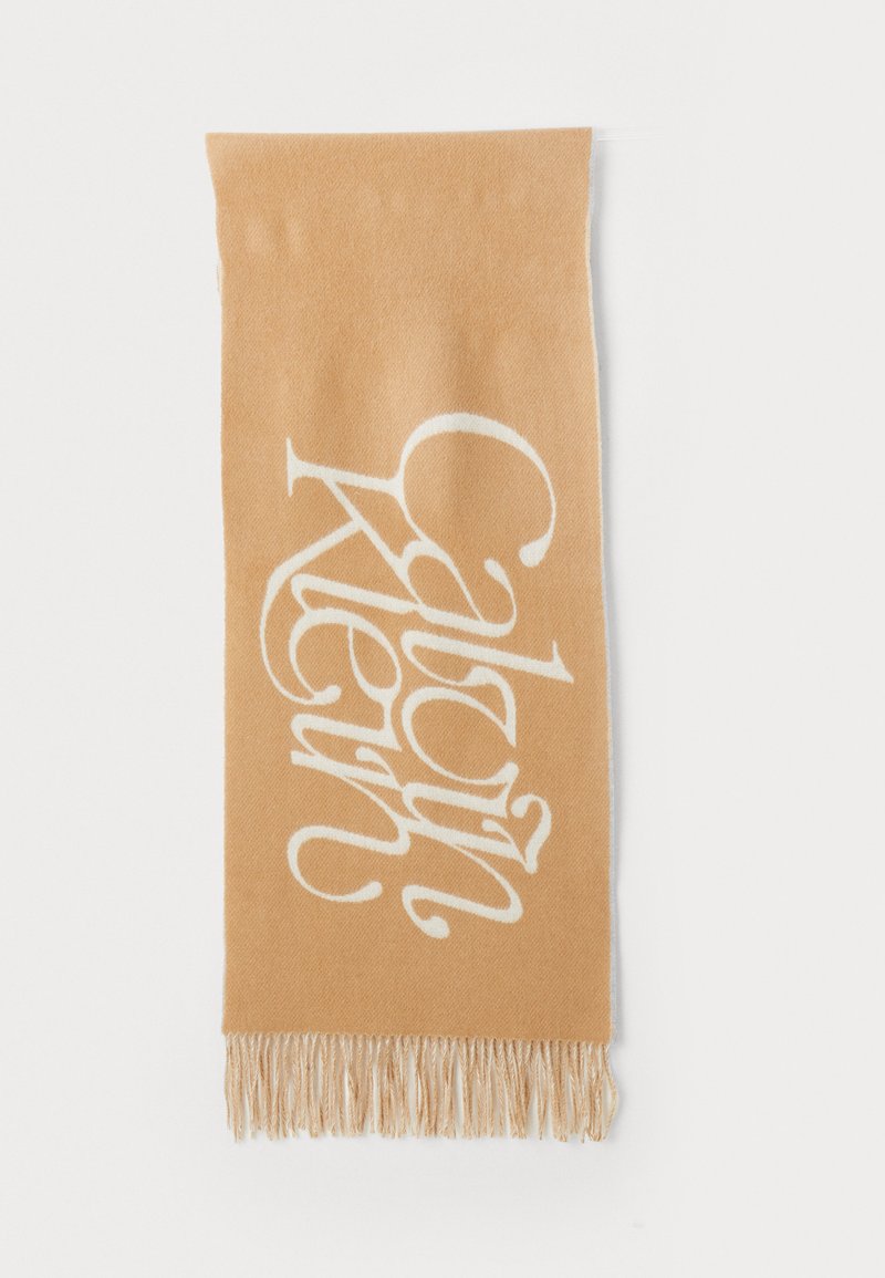 Beige tørklæde med hvid "Calvin Klein" logo design; blød tekstur, lang rektangulær form, afsluttet med frynser.