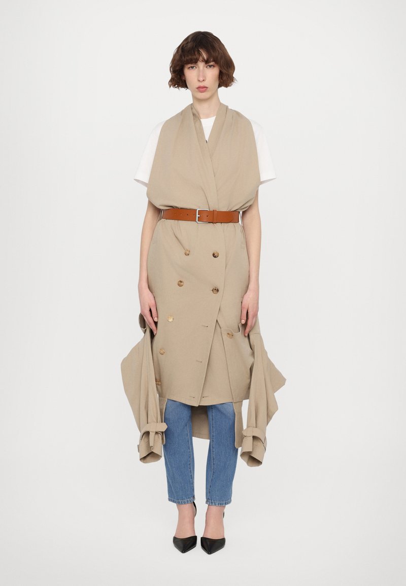 Beige, doppelt knopfverschlossener Trenchcoat mit großen Knöpfen, tailliert mit einem braunen Gürtel. Kombiniert mit blauen Jeans und schwarzen spitzen Schuhen.