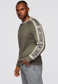 Sudadera verde oliva con cuello redondo y puños de canalé. La manga izquierda presenta un contraste en beige con texto impreso en negro que dice: "COMER DORMIR JUGAR REPETIR."