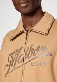 Krušņotas krāsas jaku ar rāvējslēdzēju un lielu brūnu "Malbon Golf" logotipu, rāvējslēdzēja aizdare un kontrastējoša izšuvuma apkakle. Iekļauta teksta detaļu apstrāde.