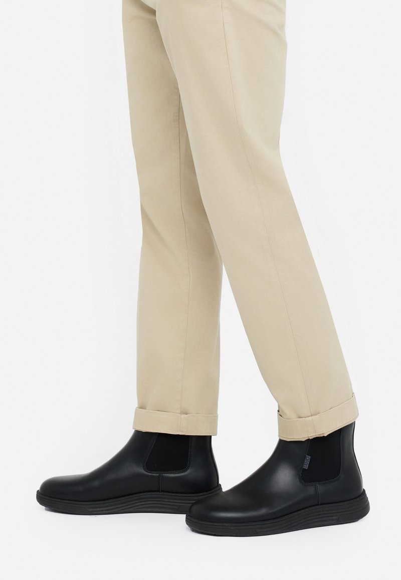 Persona con pantaloni beige arrotolati alle caviglie e stivali Chelsea neri in pelle con pannelli elastici laterali su sfondo bianco.