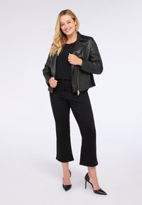 Giacca da motociclista in pelle nera con chiusura a zip, indossata sopra una maglietta nera, abbinata a pantaloni neri a cropped e tacchi a spillo neri.