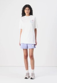Balta per didelė t-shirt su pastelinių mėlynų ir rožinių atspaudų logo ant krūtinės, derinama su šviesiai purpurinėmis šortais ir neutraliomis sportinėmis avalynėmis.