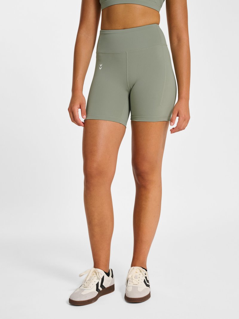 Vrouw draagt lichtgroene sportshort met hoge taille en bijpassende cropped top met witte en zwarte sneakers, staande op een witte achtergrond.