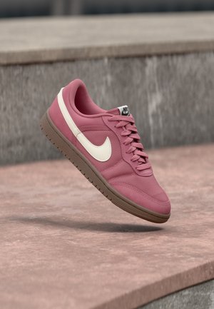 Roze Nike sneaker met witte swoosh en bruine zool, vastgelegd in de lucht boven een betonnen oppervlak met een grijze stenen achtergrond.