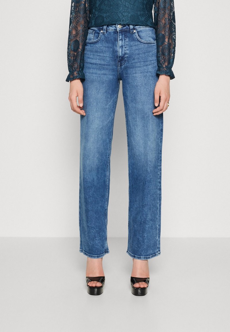 Femme portant un jean bleu taille haute à jambes droites, des talons plateformes noirs à bout ouvert et un top en dentelle vert foncé à manches longues.