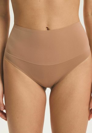 Žena nosiaca bezšvové telové tvarovacie nohavičky s vysokým pásom, zobrazujúca strednú časť tela, boky a hornú časť stehien na bielom pozadí.