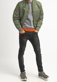 Grüne Bomberjacke, grauer Pullover mit orangefarbenem Saum, schwarze Slim-Fit-Jeans und grüne Sneakers. Die Jacke verfügt über eine Reißverschlusstasche am Arm.