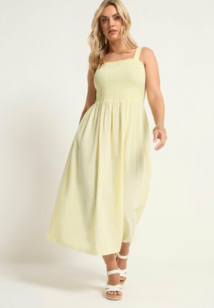 Blonde vrouw draagt een mouwloze lichtgele maxi-jurk en witte platformsandalen, lopend met een neutrale achtergrond.