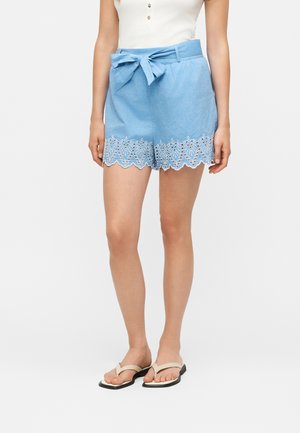 Kobieta nosząca jasnoniebieskie szorty z wiązaniem w talii, z falistą koronką z oczkami oraz beżowe płaskie sandały, stojąca na białym tle.