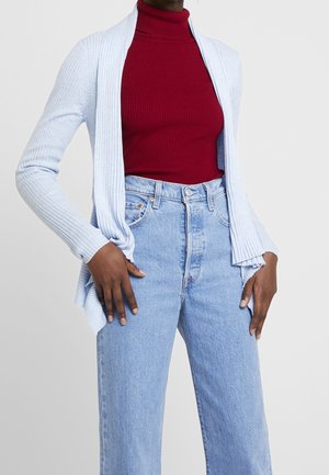 Femme portant un col roulé côtelé rouge, un cardigan ouvert côtelé bleu clair, et un jean taille haute bleu clair avec les mains dans les poches.