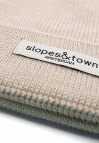 Beżowa dziana czapka z prążkowanym wzorem. Posiada biały metkę naszytą, na której widnieje napis "slopes&town AMSTERDAM" w czarnym foncie.