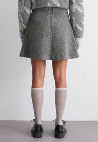 Persoon in een grijze flare rok, een patronen trui, glinsterende doorschijnende kniekousen en zwarte veterschoenen, staand op een grijze vloer.