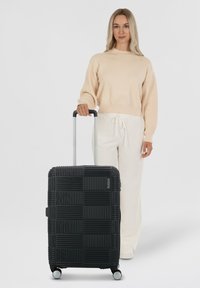 Maleta rígida negra con un patrón cuadrado texturizado, mango telescópico plateado y ruedas lisas. Se mantiene en posición vertical al lado de una persona vestida con ropa clara.