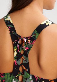 Blumengemusteter Stoff mit schwarzem Hintergrund zeigt ein Spitzenrücken-Design. Die Träger sind mit metallischen Ösen und bunten Schnürsenkeln verziert.