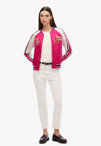 Veste en bomber rose avec des fleurs brodées, manches blanches, détails rayés, t-shirt blanc, pantalon blanc et mocassins foncés.