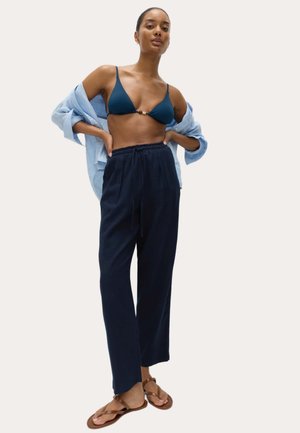 Kvinde iført navy bikini top, lyseblå åben skjorte, løse mørkeblå bukser og brune sandaler, stående med hænderne på hofterne mod en ensfarvet baggrund.