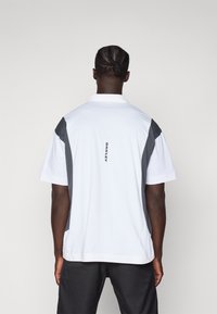 Kortermet polo-skjorte i hvit, med grå skulderpaneler og logo på baksiden. Stoffet ser glatt ut med et klassisk kragedesign.