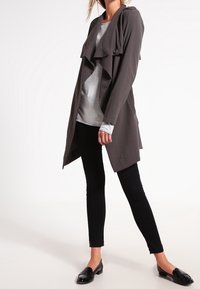 Giacca lunga grigia, leggera, con davanti drappeggiato e dettaglio con bottoni, indossata sopra una maglietta grigia chiara e pantaloni neri slim, abbinata a mocassini neri.